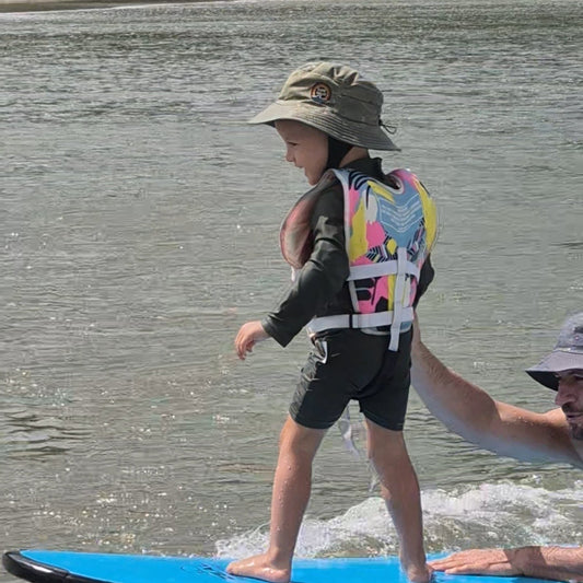 A young child stands on a blue surfboard in shallow water, wearing a multicolor life vest, black rash guard, and a surf hat helmet with his dad helping him learn surfing
